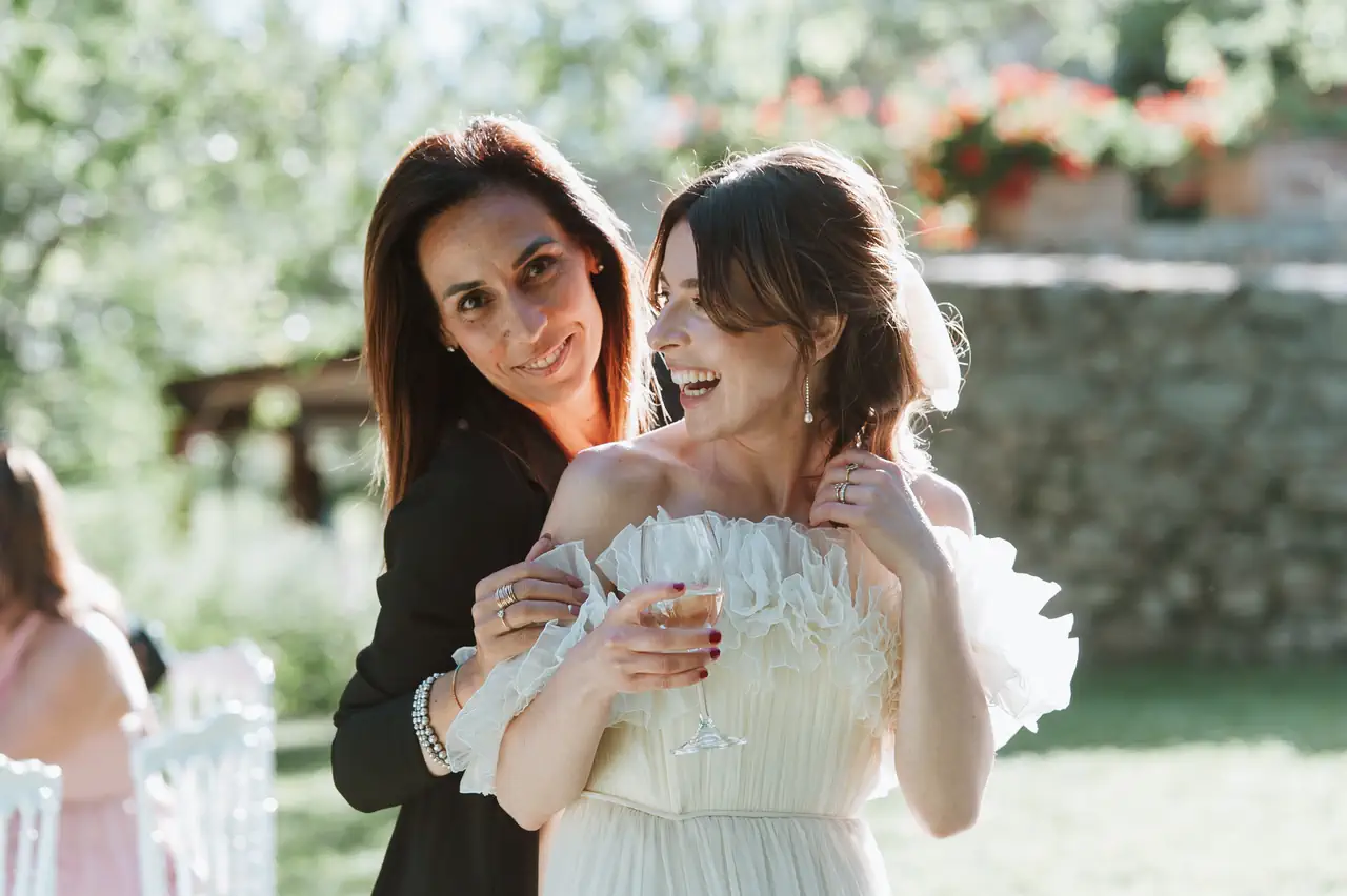 Contact. Two women in the open, the bride in a white dress holding a glass of wine and smiling, simona celani standing behind her in a dark dress with her hands on her shoulders.