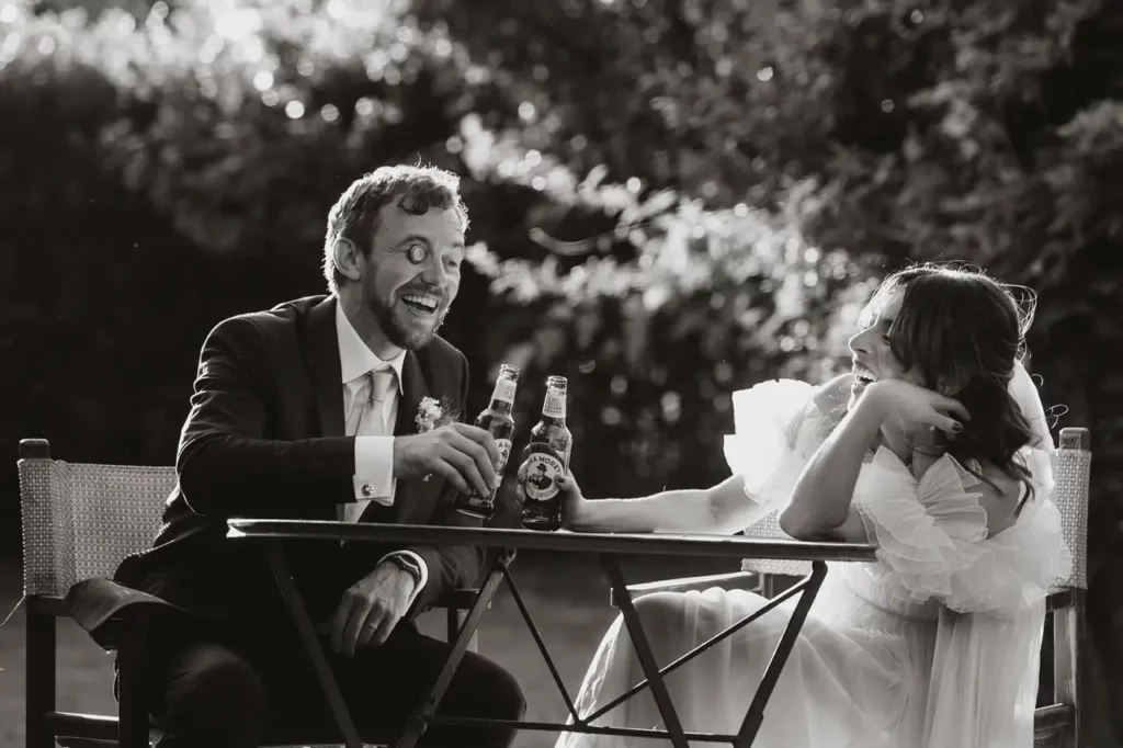 Black-and-white photo of a bride and groom sitting at an outdoor table, sharing a laugh while holding bottles of beer.