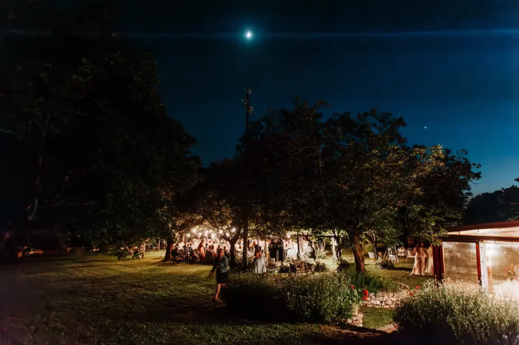 A night scene of an outdoor event at the groundhog village, with lights illuminating people under the trees and a bright moon shining in the sky.