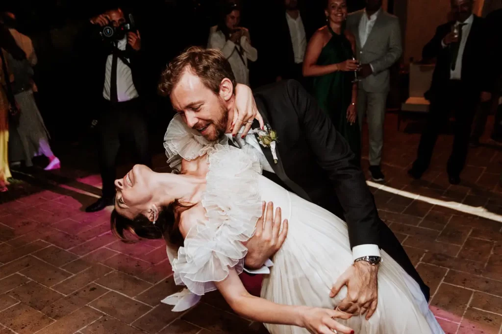 A groom holds the bride during the dance at their wedding reception. People in formal attire appear in the background, some taking pictures and watching.