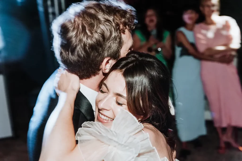 A woman and a man hug each other tightly as they dance. The bride has a joyful expression. Several people are in the background observing their dance.