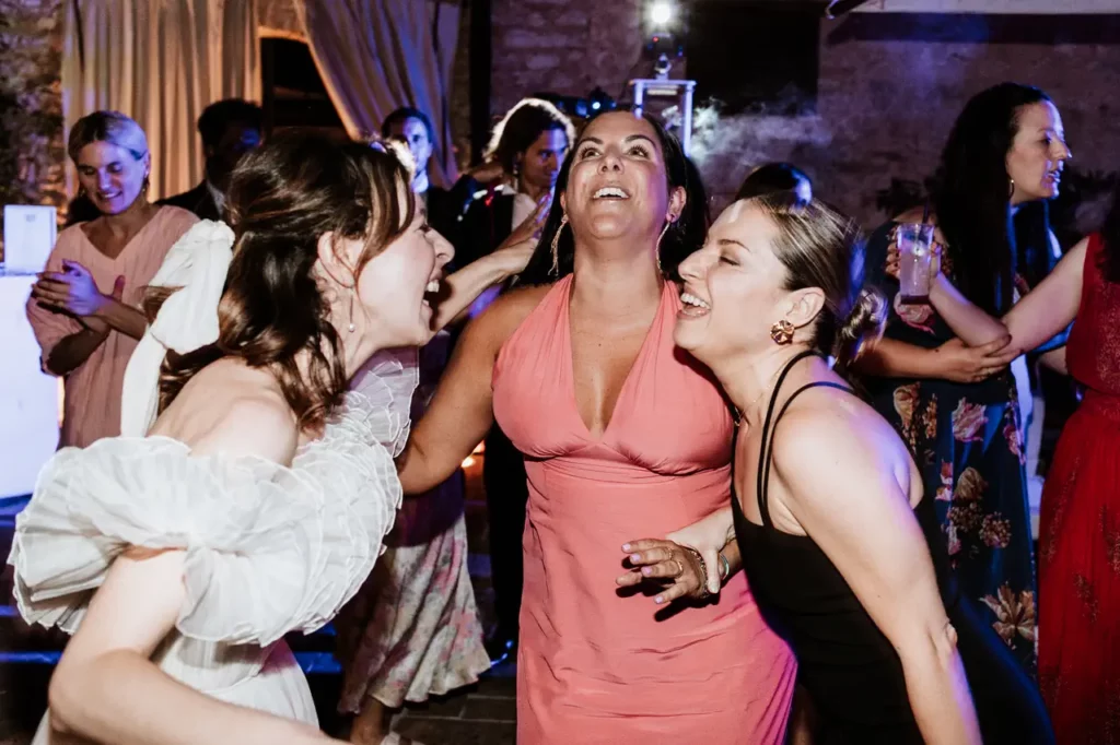 Three women laugh and hug each other on a dance floor. One is wearing a white dress, another a pink dress, and the third a black dress. Other people are visible in the background.