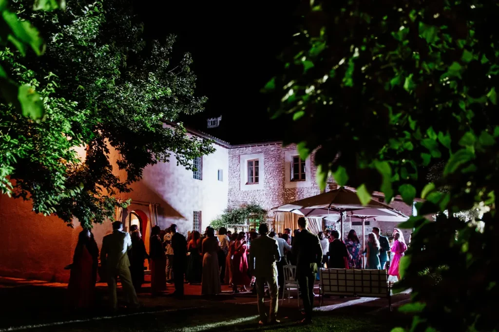 Night scene of people gathered at the borgo della marmotta, socializing under trees and umbrellas. The scene is lively with a mix of light and shadow.