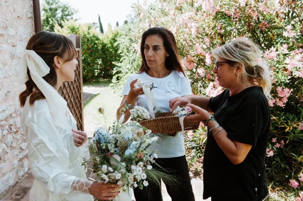 Three women are standing outdoors: the bride, holding a bouquet, next to a woman dressed in black (the florist) delivering items from a basket to another woman wearing a white t-shirt (simona celani). They are leaning against a stone wall and surrounded by greenery.