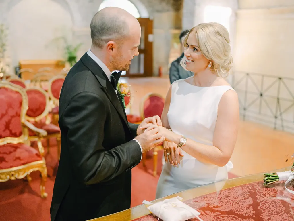 The bride and groom exchange rings in an elegantly decorated interior with red chairs and a table with gold accents in the background. The bride wears a white suit and the groom a black suit.