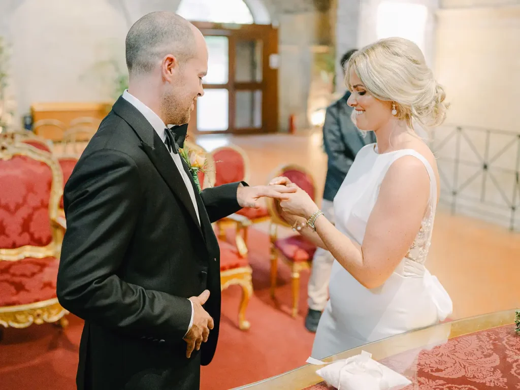 The bride and groom exchange rings in an elegant hall with red chairs and elaborate decorations. The bride wears a white suit; the groom wears a black suit.