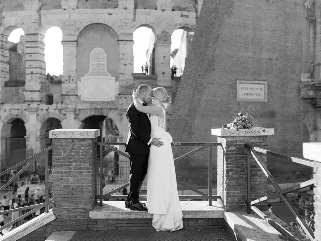 A formally dressed couple kisses on a platform in front of ancient ruins.