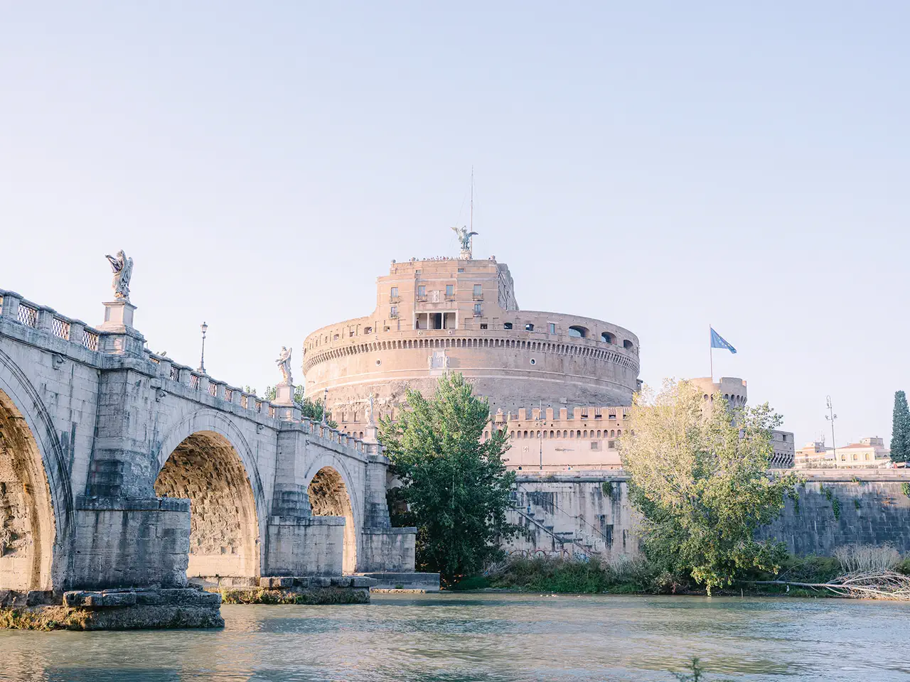 The historic stone bridge with arches crosses the Tiber in front of the imposing Castel Sant'Angelo, surmounted by statues, against a clear sky with greenery visible from the water.