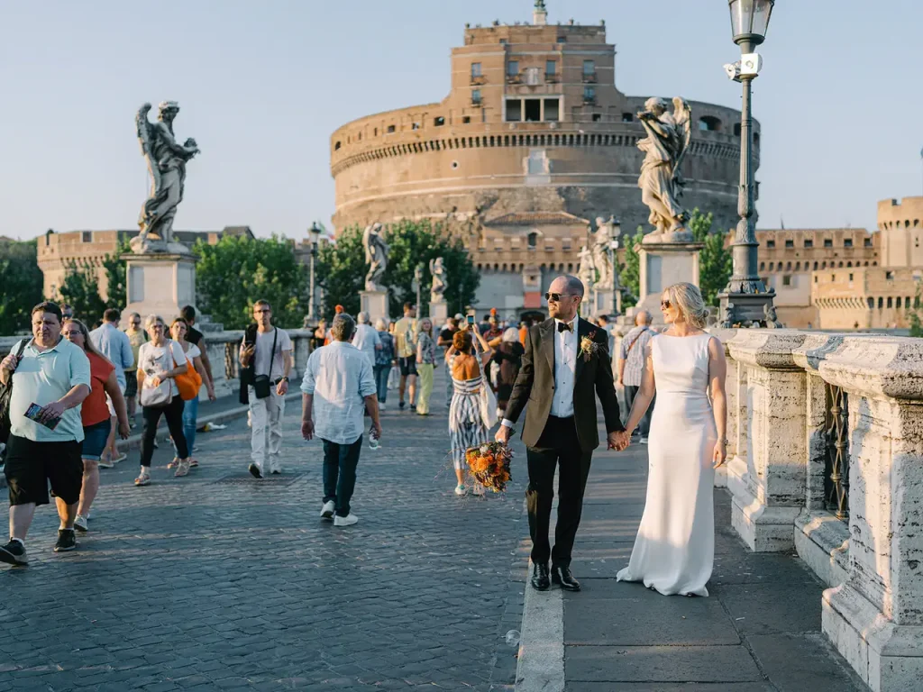 A couple in formal dress walk hand in hand across a busy bridge, with tourists and statues nearby. In the background stands the imposing castel sant'angelo, a testimony to history.