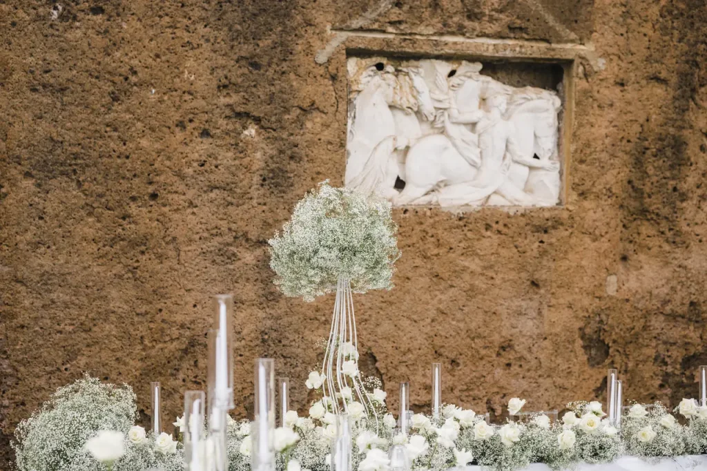 Floral arrangement with white and green flowers in front of an ancient stone relief depicting a historical scene carved on a brown textured wall. Transparent cylindrical decorations are visible.