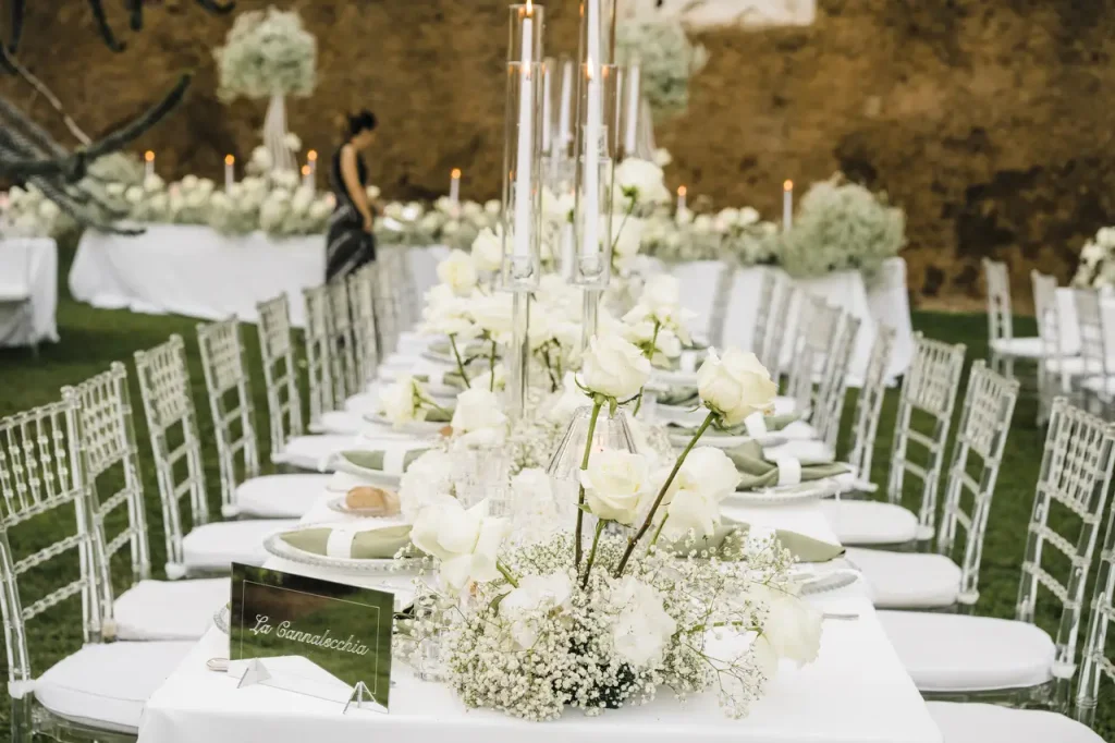 A long banquet table is set up for a formal event with white floral centerpieces, candles, and well-placed place settings. A person in the background appears to be setting up or inspecting the area.