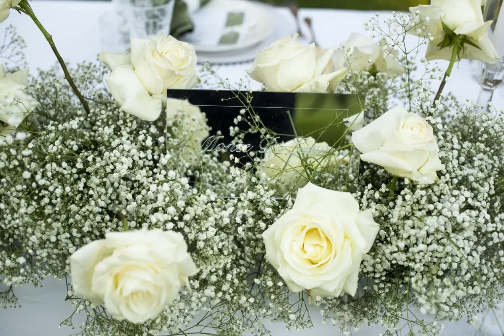 Close-up of a floral arrangement with white roses and gypsophila surrounding a small rectangular mirror on a table.