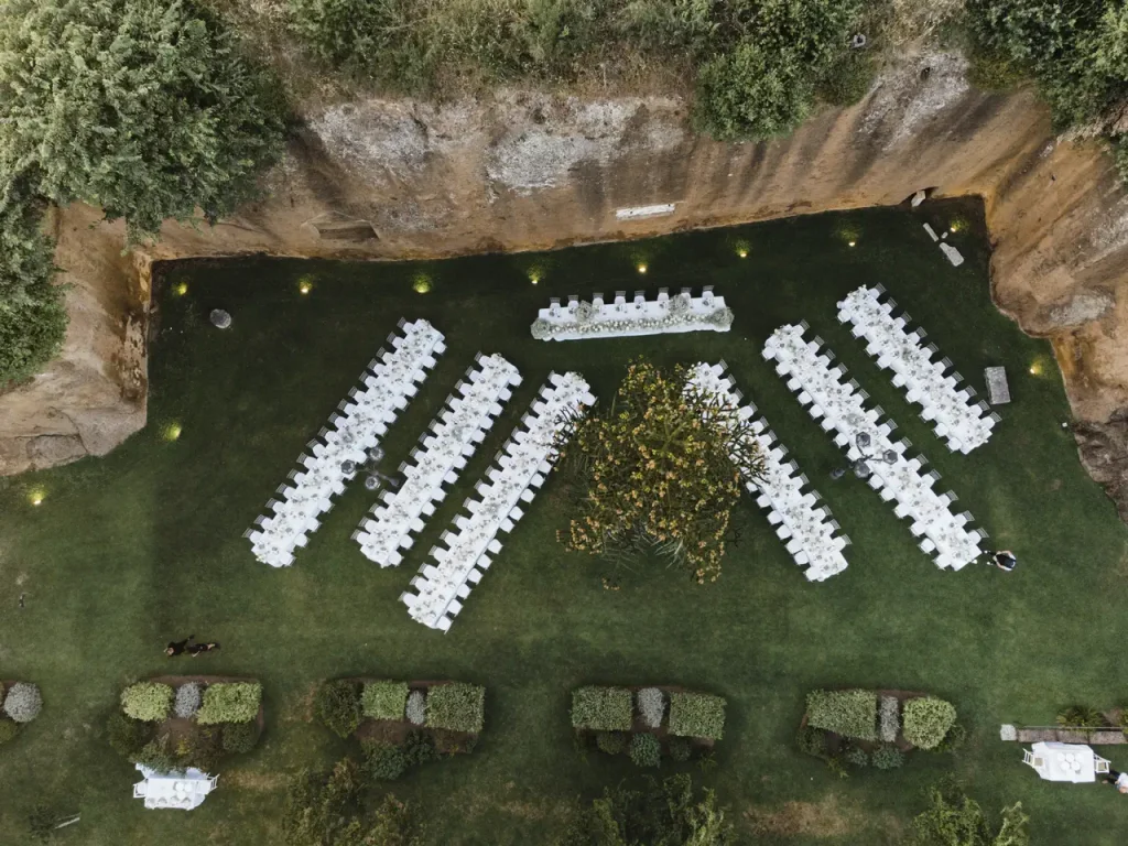 Aerial view of an outdoor wedding reception set up on a grassy area at the antica flaminia residence, with long white tables and chairs arranged in rows, surrounded by greenery and a rock wall.