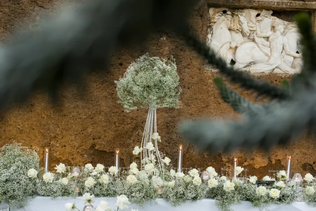 A decorative table set with white flowers and candles is depicted against the background of a stone wall with intricate carvings. Pine branches partially frame the foreground.