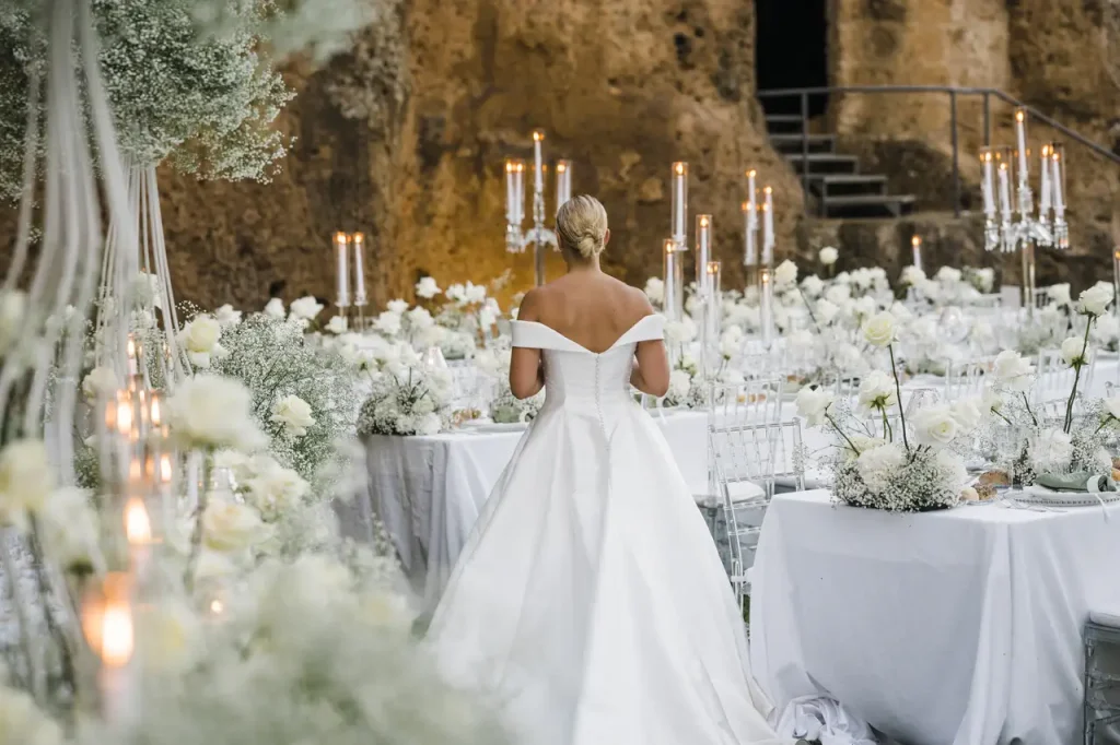 A bride in a white gown with bare shoulders stands in front of elegantly set tables decorated with white flowers, candles and greenery, with a rocky backdrop behind her.