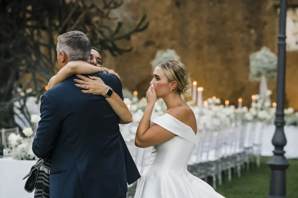 A bride in a white dress stands with her hand over her face as the groom is embraced by simona celani during an outdoor event with long tables and white decorations.