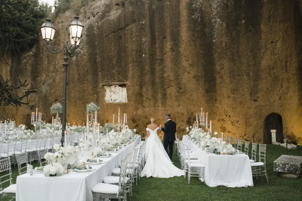 A bride and groom walk between decorated tables set up for an outdoor wedding reception with a rock wall in the background.