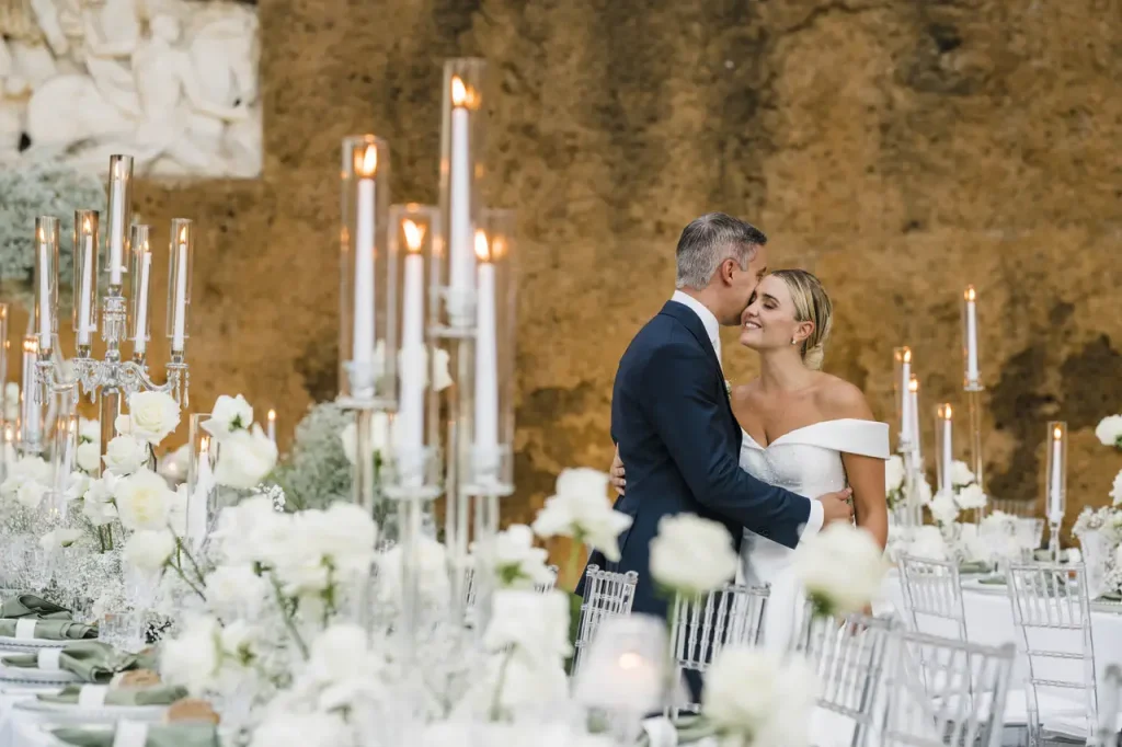 A couple embraces near a table elegantly set with white roses and tall candle holders against a rustic stone background.