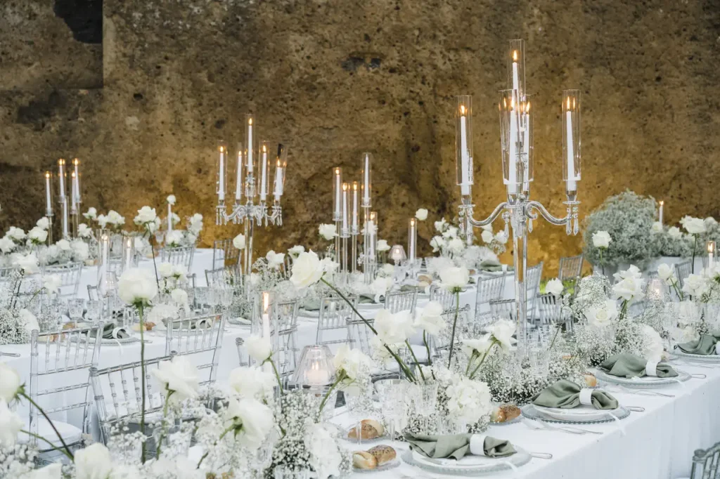 A formal dining room with white floral arrangements, white tablecloths, candles in candelabras, and place settings on rectangular tables against a rustic stone background.