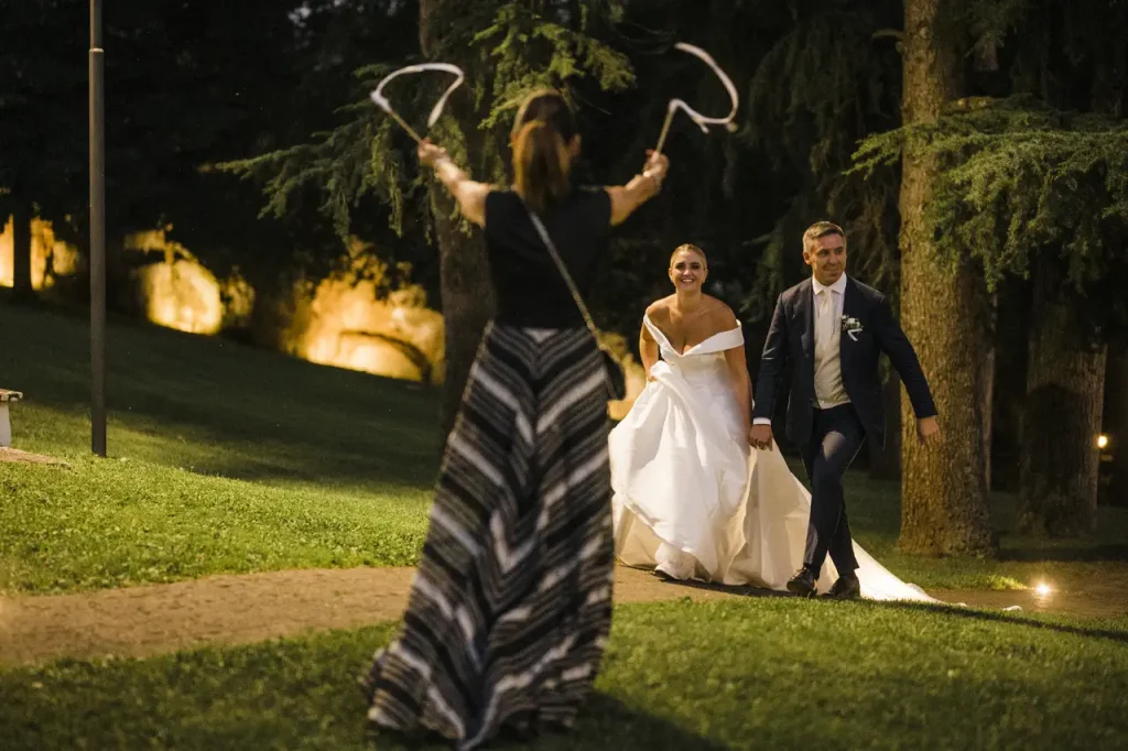 The bride and groom walk on the grass under the direction of wedding planner simona celani, with trees and lights in the background.