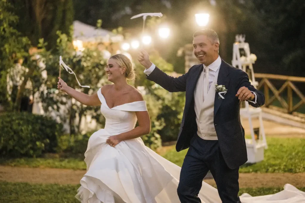 A bride and groom in formal attire joyfully wave streamers outdoors at night, illuminated by small lights. The bride lifts her dress with one hand.
