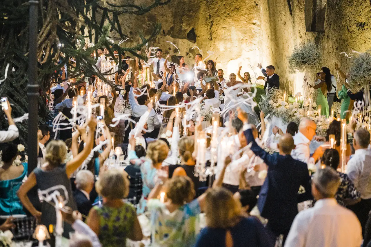 People celebrating with raised hands and streamers in a dimly lit, crowded indoor space with rough stone walls.