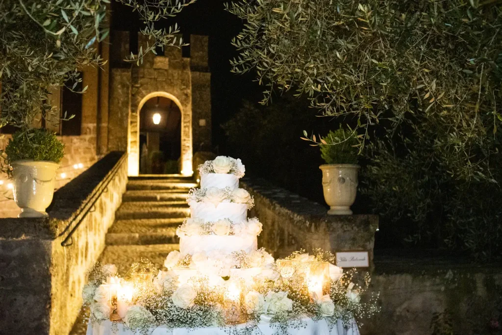 An outdoor night scene with a multi-tiered white wedding cake surrounded by lit candles on a table, positioned near a building with stone steps and lighted entrance.