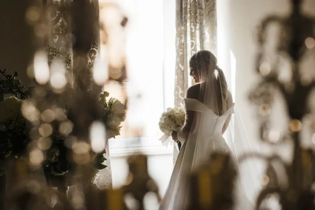 A bride in a white dress and veil stands inside near a window holding a bouquet of flowers, with decorative details and soft lighting in the background.