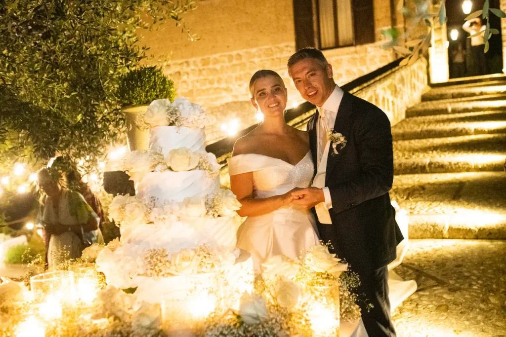 A bride and groom stand in front of a multi-tiered white wedding cake, smiling for a photo during a nighttime outdoor reception adorned with lights.