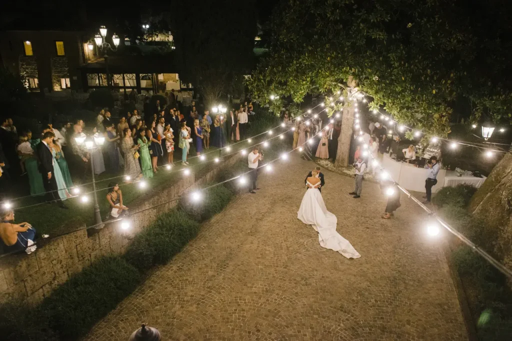 A married couple dances under string lights surrounded by guests during a nighttime outdoor event.