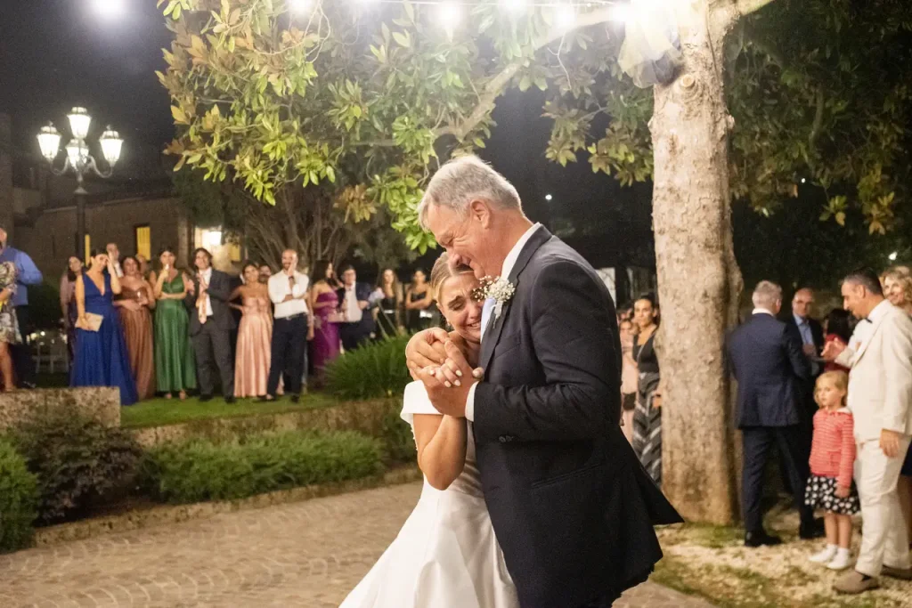 A bride and her father dance together outdoors during a nighttime wedding reception. Guests stand nearby, watching and celebrating.