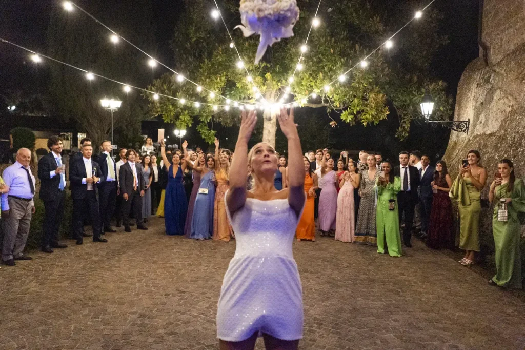 A woman in a white dress throws a bouquet of flowers in the air during an outdoor evening celebration in front of a crowd of people watching.