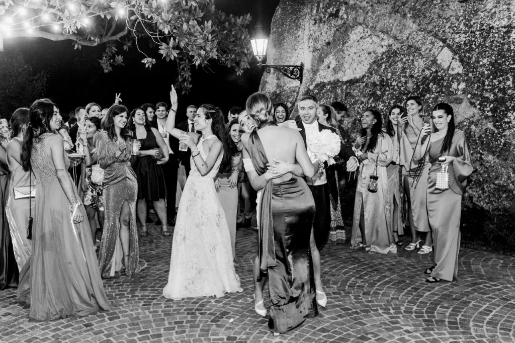 Black and white photo of the wedding bouquet toss. Gathered outside is a group of women in elegant dresses; one woman has caught the bouquet and is hugging the bride, while others are rejoicing.
