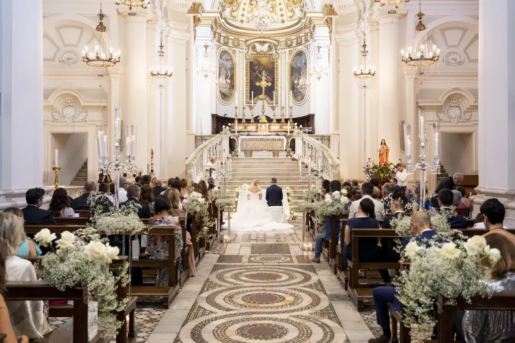 The bride and groom kneel before the altar during a wedding ceremony at the cathedral of santa maria maggiore in civita castellana, a large and ornate church filled with guests. The church features white columns, chandeliers and intricate ceiling details.