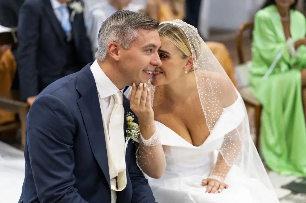 A bride in a white dress and veil bends down to whisper to the groom in a dark suit and tie during the wedding ceremony.