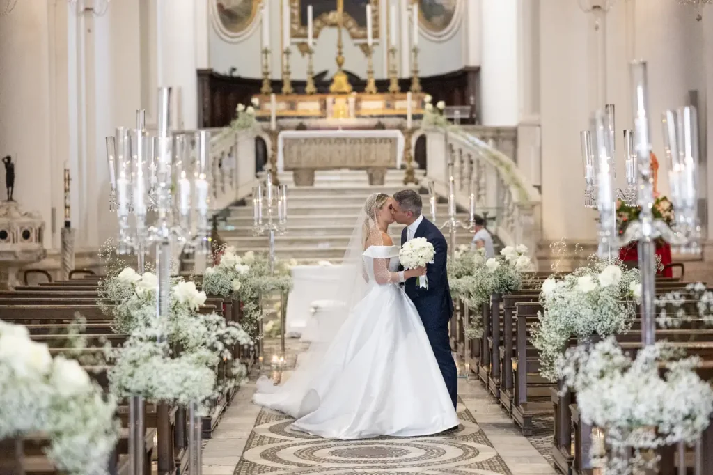 A couple in wedding gowns share a kiss at the altar of an ornate church, with white floral arrangements and candle holders along the aisle.