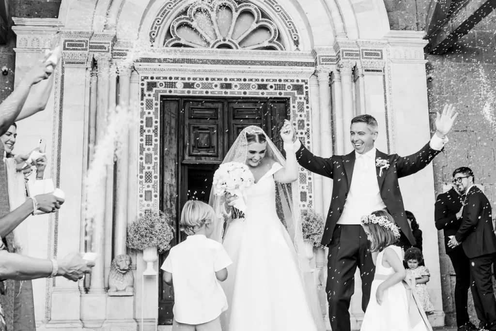 A newly married couple walks out of a church holding hands, smiling and celebrating. Guests, including children, stand nearby and shower them with confetti. The scene is joyous and festive.