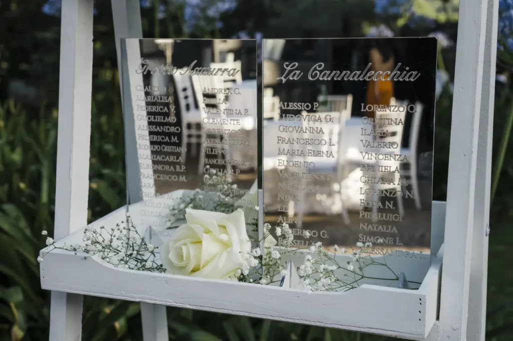 Two mirrored glass chalkboards display names and are adorned with white roses and baby's breath, placed on a white shelf.