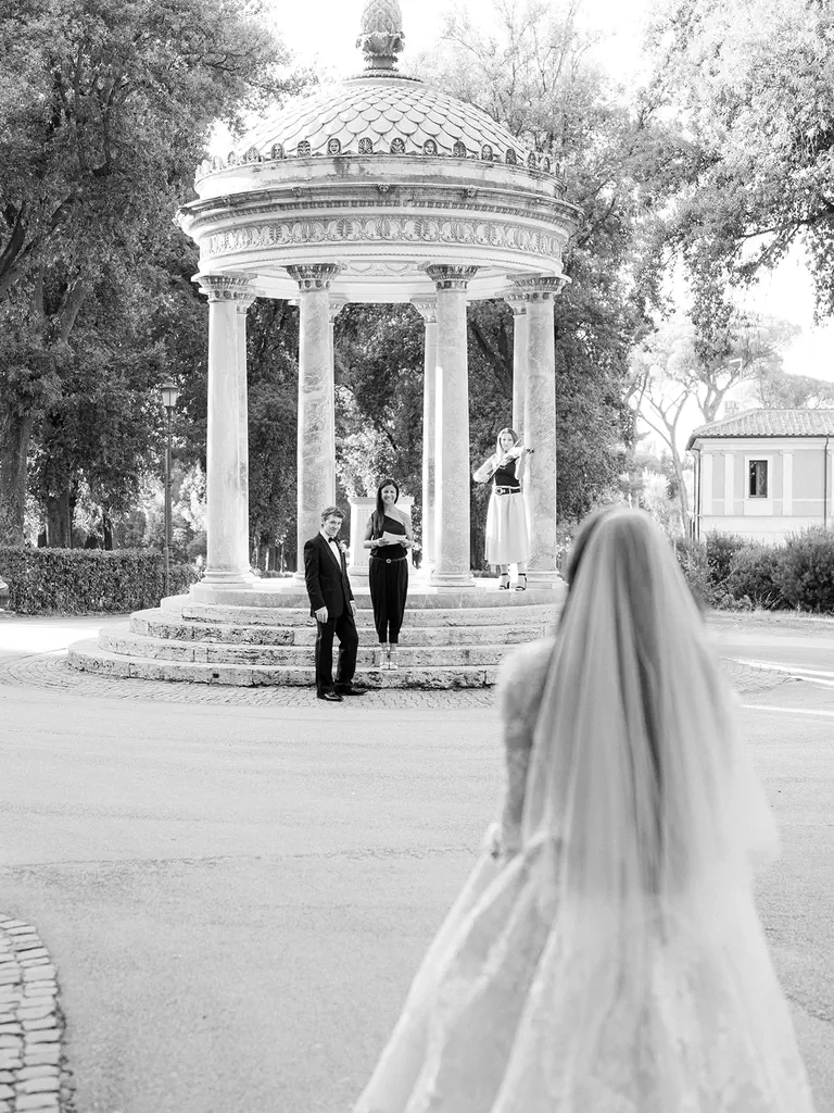 Jordan e kelly, elopement a roma Una sposa si dirige verso il tempietto di diana dove stanno uno sposo, il cerimoniere e la violinista. La scena è ambientata all'aperto con alberi ed edifici sullo sfondo. L'immagine è in bianco e nero.