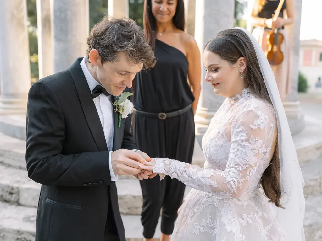 A groom dressed in black puts a ring on the bride's hand while a woman dressed in black stands behind them. The bride wears a lace wedding dress with a veil.