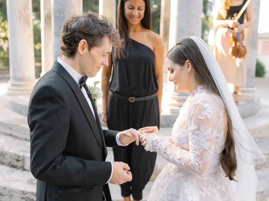 A groom dressed in black places a ring on the bride's finger during an outdoor wedding ceremony, with an officiant and a violinist in the background.