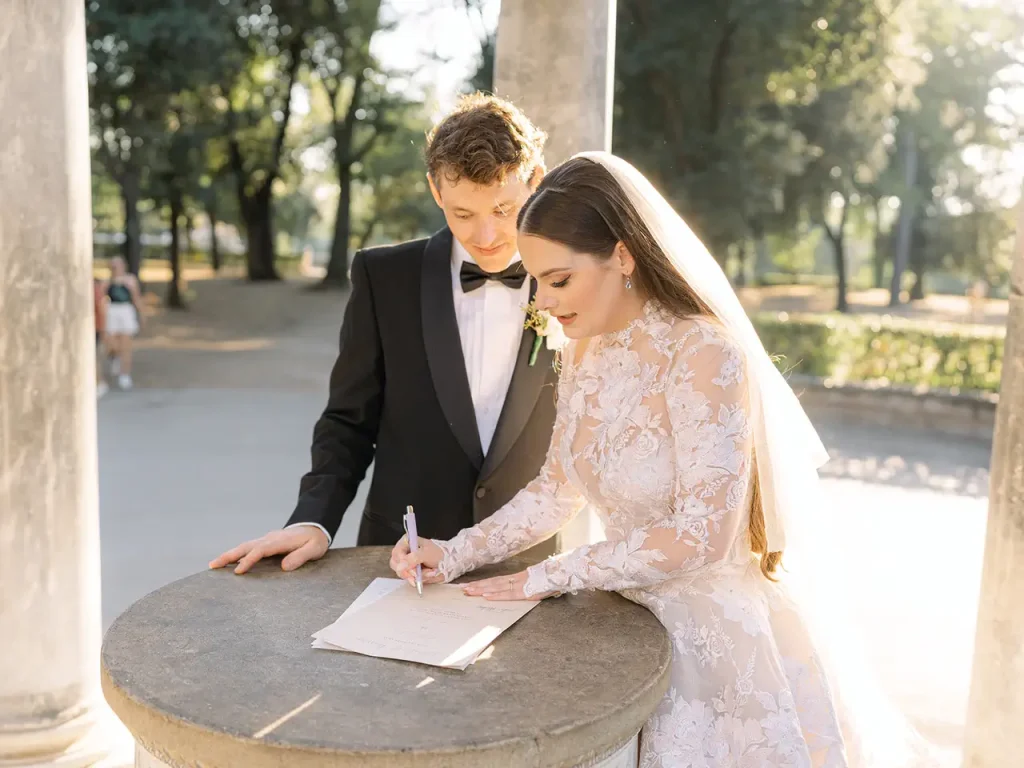 A bride and groom sign a document on a stone pedestal in an outdoor setting. The bride wears a white lace dress and the groom a black tuxedo. They seem focused on the task at hand.