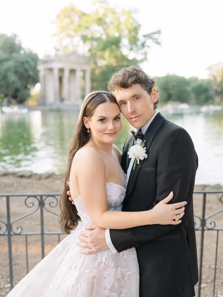 Jordan and kelly, elopement in rome A couple dressed in wedding attire pose for an outdoor photo by a lake, with a classical building in the background. The bride wears a white suit, while the groom wears a black suit with a flower in the buttonhole.