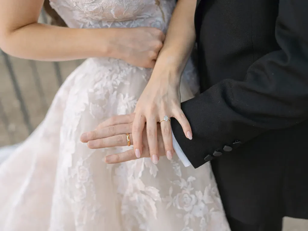 A close-up of a couple showing wedding rings. The woman is wearing a lace wedding dress and the man is wearing a black suit. Their hands are intertwined.