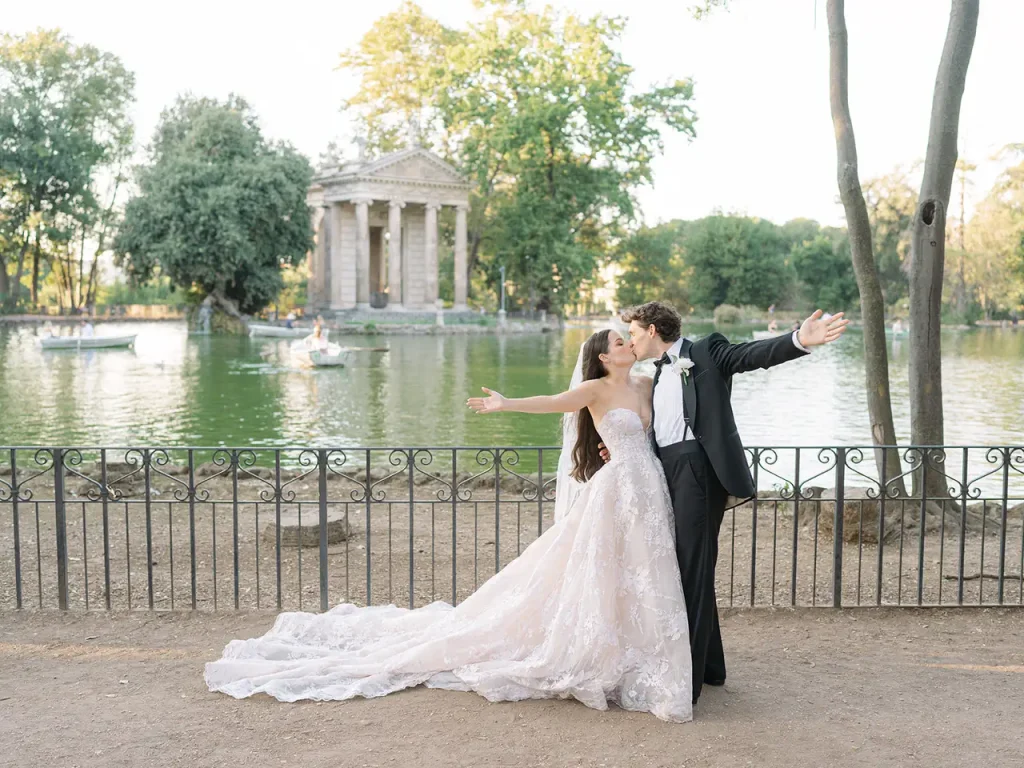 Kelly and jordan's elopement: a couple in wedding gowns embrace with outstretched arms while standing in front of a lake and the classical temple of aesculapius at villa borghese.