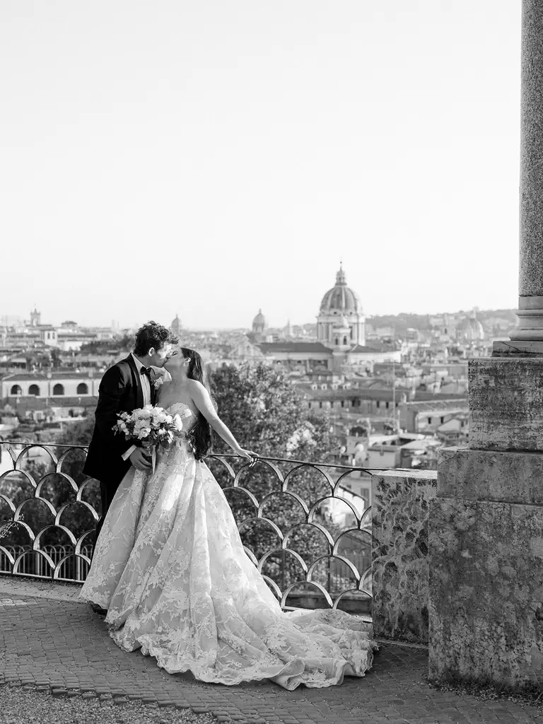 A bride and groom kiss in front of a viewpoint featuring the rome skyline of domed buildings and historic architecture. The bride holds a bouquet and wears a lace dress. The image is beautifully captured in black and white.