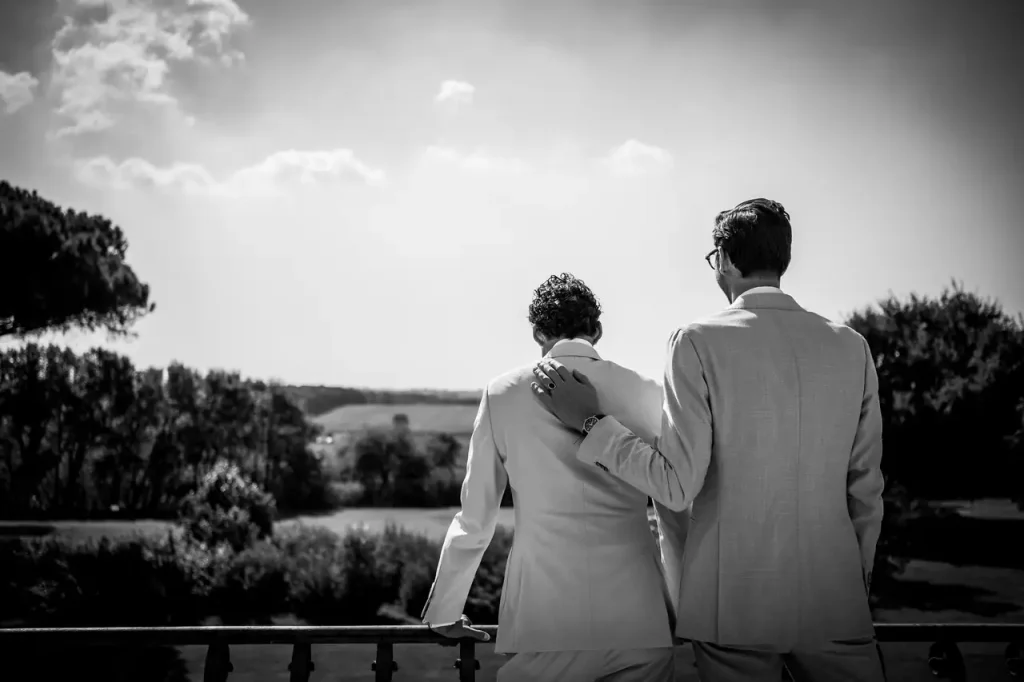 Two men in suits are standing on a balcony of villa alberico, overlooking a picturesque landscape. One person has his hand on the other's shoulder. The image is in black and white.