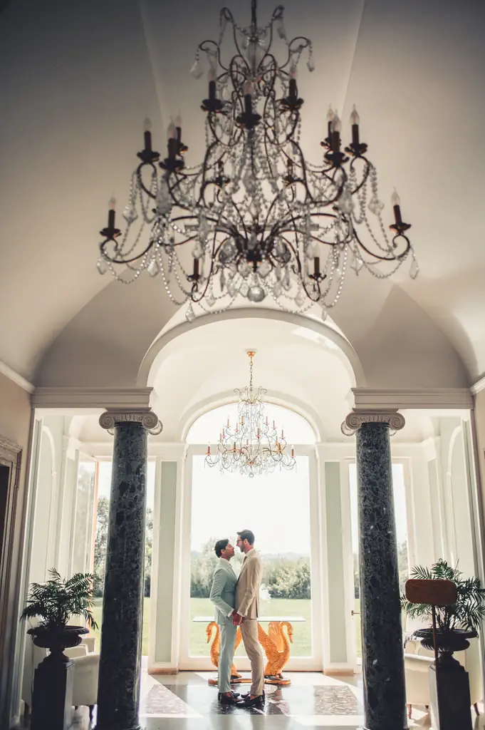 Marco diederik wedding villa alberico Two men are standing, facing each other, in an elegantly decorated room with large windows, marble columns, and a crystal chandelier above their heads.