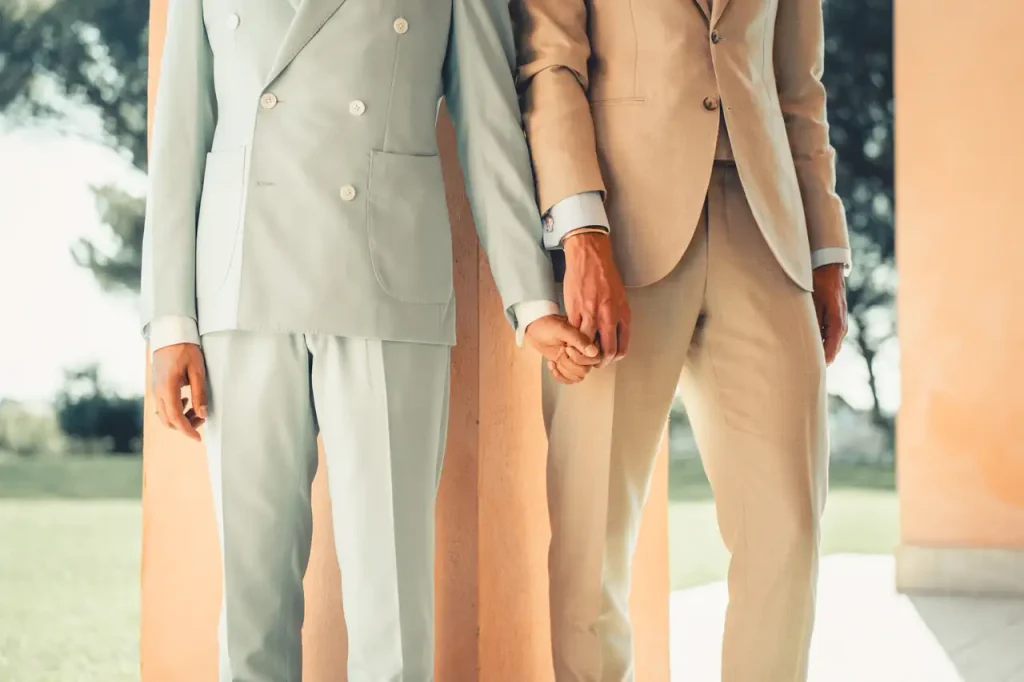 Two people wearing light-colored clothing are standing next to each other, holding hands, with an outdoor environment in the background. Their faces are not visible.