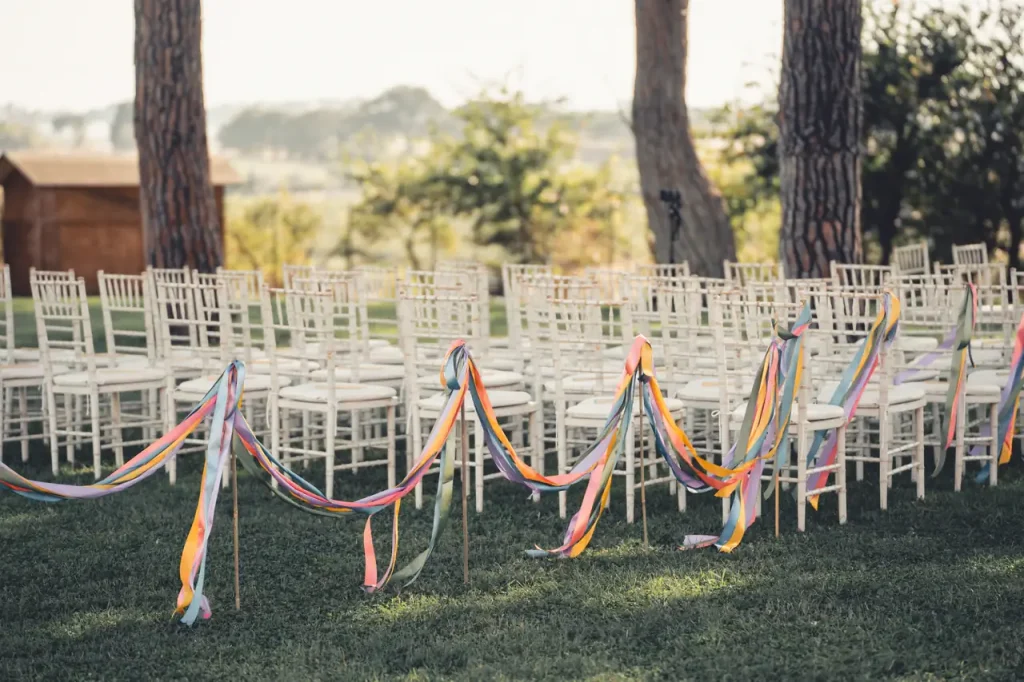 Outdoor wedding ceremony setup with white chairs arranged in rows on a lawn, decorated with colorful ribbons strung between poles. Trees and a wooden structure are visible in the background.